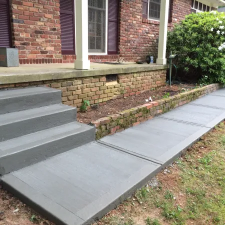 Freshly poured gray concrete steps and walkway leading to a red brick house with green landscaping.
