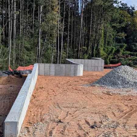 Concrete foundation walls and gravel pile at a construction site surrounded by trees and orange safety fencing.