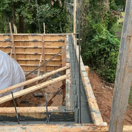Freshly poured concrete foundation wall with wooden forms and rebar in a forested outdoor construction site.