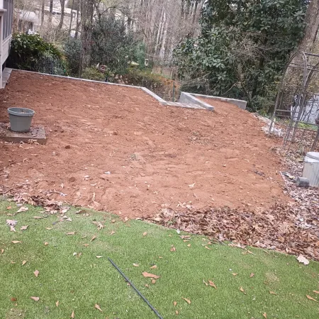 Cleared backyard area with fresh soil and surrounding fallen leaves near green grass and garden pots.