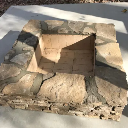 Square stone fire pit set on a concrete patio surrounded by autumn leaves in a woodland setting.