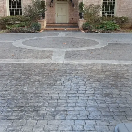 Stamped concrete driveway with circular and linear patterns leading to a brick house entrance with shrubs.