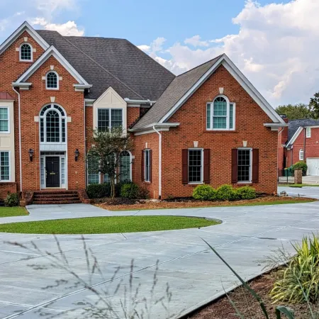 Large brick two-story house with multiple windows, arched design, and a wide concrete driveway under a cloudy sky