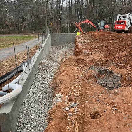 Construction site with excavator and compact loader working on soil and concrete foundation walls under trees.