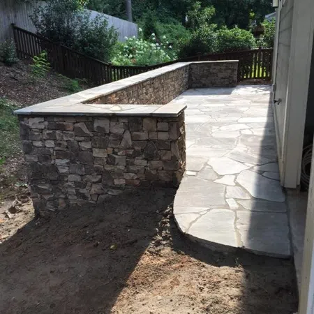Stone retaining wall with a concrete patio area next to a house surrounded by garden and trees.
