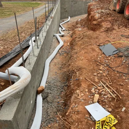 Drainage pipes installed alongside concrete foundation walls at a residential construction site with machinery and caution tape.