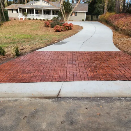 Curved concrete driveway with red brick-patterned section leading to a house surrounded by trees and landscaping.