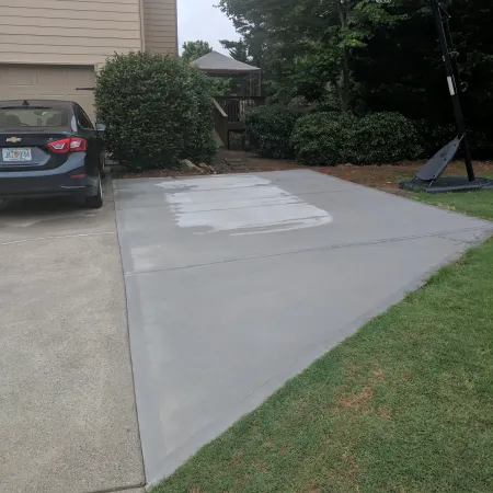 Freshly poured concrete driveway beside a car and basketball hoop, surrounded by greenery.