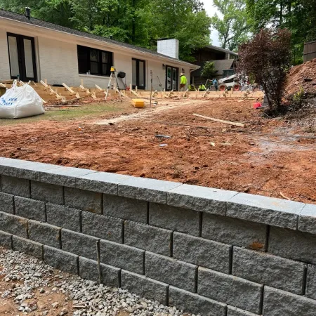 Stone retaining wall with fresh gravel base and construction workers building a wooden frame near a brick house.
