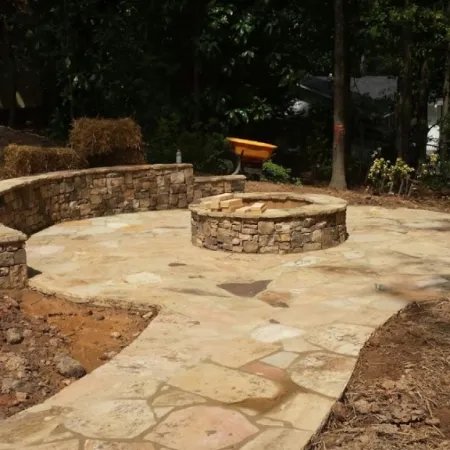 Beautiful stone patio with circular fire pit surrounded by lush green trees and natural landscaping.