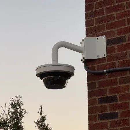 White dome security camera mounted on a red brick wall with trees and clear sky background.