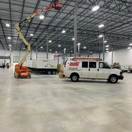 Interior of a spacious industrial warehouse with a white utility van and an orange boom lift raised to the ceiling.