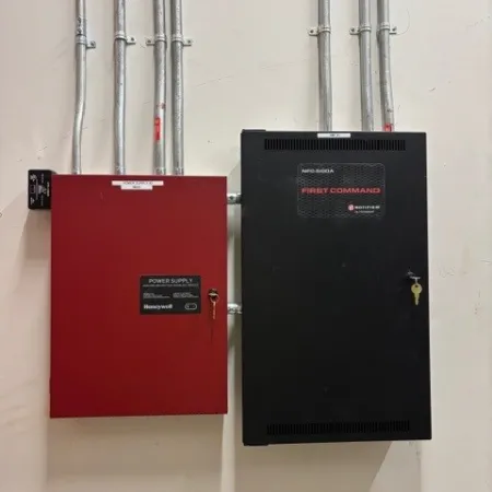 Red and black metal control panels mounted on a white wall with multiple electrical conduits above them.