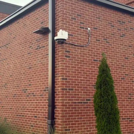 Security camera mounted on red brick building corner with black downspout and green shrubs nearby