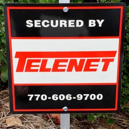 Security sign for Telenet with contact number displayed in garden area with greenery and dry leaves.