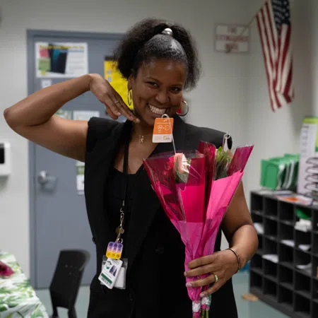 Smiling woman holding a bouquet of pink-wrapped flowers posing happily in an office setting with an American flag.
