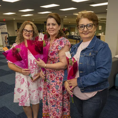 Three smiling women holding pink rose bouquets indoors, wearing floral and denim outfits.