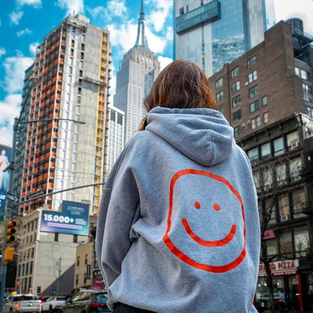Person wearing grey hoodie with orange smiley face, standing in city with skyscrapers and blue sky.