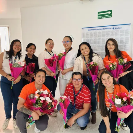 Group of smiling healthcare workers holding pink flower bouquets in a bright hospital hallway.