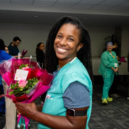 Smiling nurse in scrubs holding a bouquet of flowers during a hospital staff appreciation event indoors.