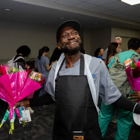 Smiling man in apron holding pink-wrapped flower bouquets in a crowded indoor room with diverse people.