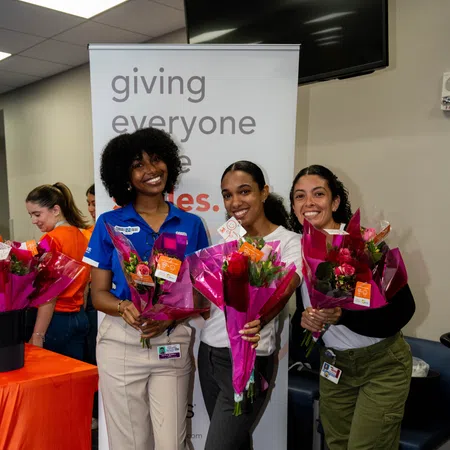 Three smiling women holding flower bouquets standing in front of a banner at an indoor event.
