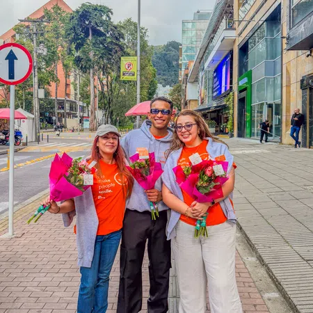 Three smiling people holding bouquets of flowers standing on a city sidewalk wearing orange shirts and gray sweaters.