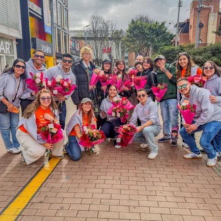Group of diverse young people posing outdoors holding pink flower bouquets on a city sidewalk.