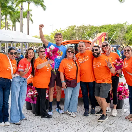 Group of smiling people wearing orange shirts posing outdoors with flowers and palm trees in background