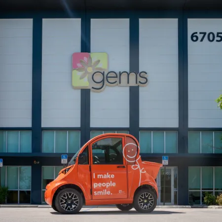 Orange compact car with smiling face and text parked in front of Gems company building with address 6705.