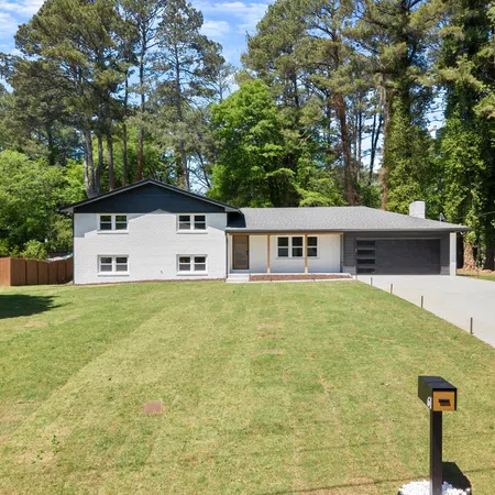 Modern two-story white house with a large green lawn, concrete driveway, and surrounding tall trees under blue sky