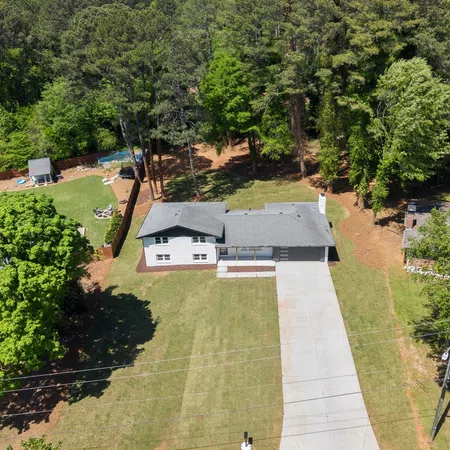 Aerial view of a single-story house with gray roof, large driveway, and surrounding green trees and lawn.