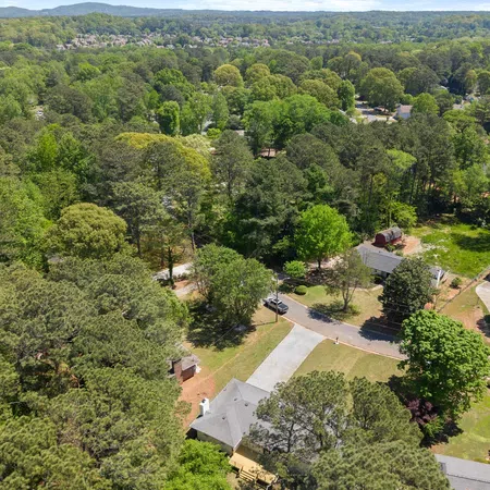 Aerial view of suburban neighborhood surrounded by dense green trees and distant hills under clear sky.
