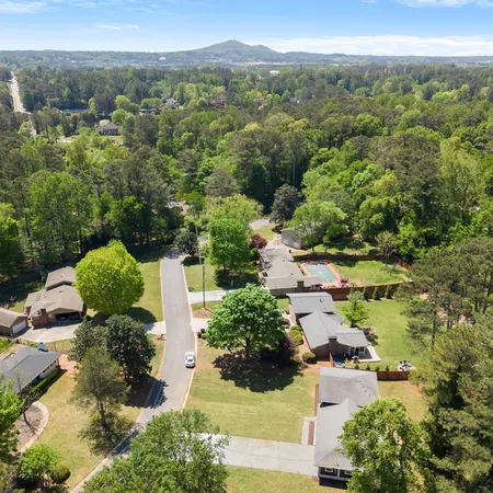 Aerial view of a suburban neighborhood with houses, green trees, lawns, and a distant mountain under a clear blue sky.