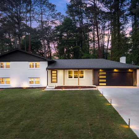 Modern two-story white brick house with black roof and garage at dusk surrounded by tall pine trees and lit driveway.