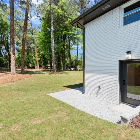 Backyard view with a green lawn, tall trees, and the side of a white brick house with a door and window.