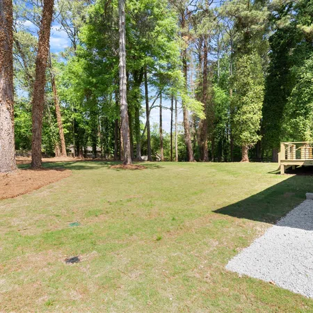 Spacious backyard with green grass, tall trees, and a deck attached to a house on a sunny day