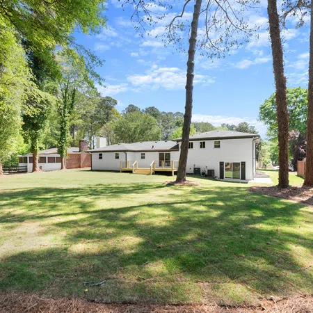 Spacious backyard with green lawn, tall trees, and a white house under a blue sky with scattered clouds.