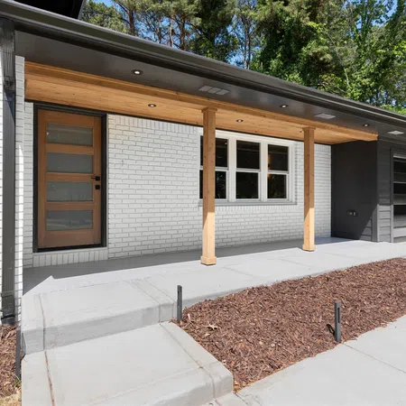 Modern house exterior with white brick walls, wooden front door, porch with wooden columns, and concrete walkway.