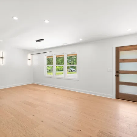 Bright modern empty room with light wood flooring, white walls, large window, dark cabinets, and wooden door.