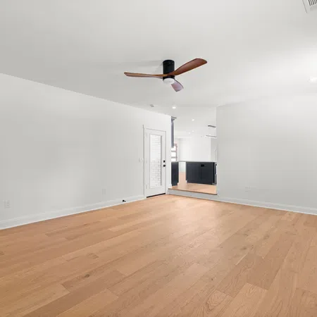 Empty modern living room with light wood flooring, white walls, ceiling fan, and natural light from windows.