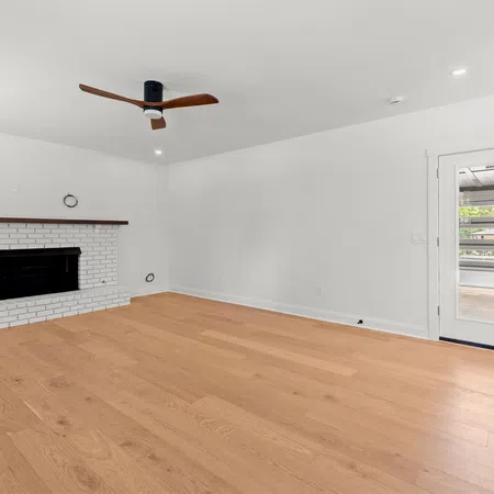 Empty modern living room with light hardwood floors, white walls, brick fireplace, ceiling fan, and glass door.