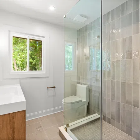 Modern bathroom with glass shower enclosure, wood vanity, wall sconce lighting, and window showing green trees.