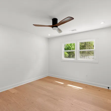 Empty bright room with white walls, wood flooring, ceiling fan, and double window showing greenery outside.