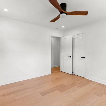 Empty modern bedroom with white walls, light wood flooring, ceiling fan, and two white doors open.