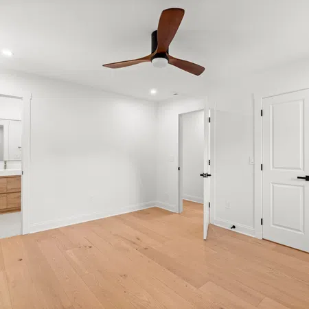 Empty modern bedroom with wooden floor, white walls, ceiling fan, and doors leading to bathroom and closet