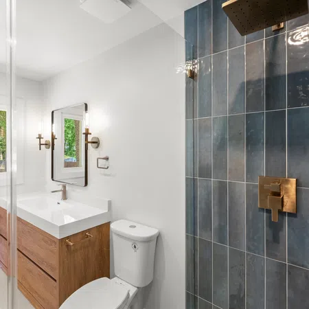 Modern bathroom with wooden vanity, white sink, toilet, large window, and dark blue tiled shower with brass fixtures