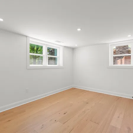 Bright empty room with white walls, light wood flooring, two windows, and recessed ceiling lights.