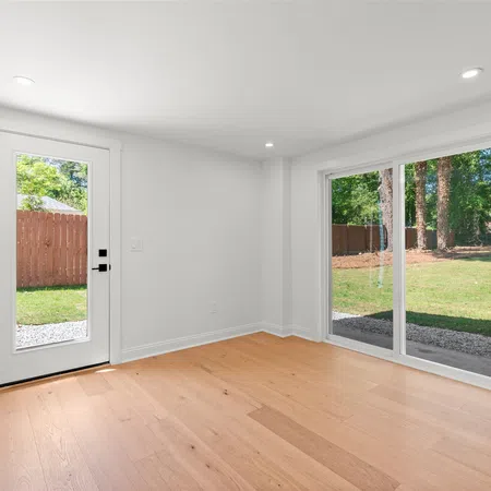 Bright empty room with light wood flooring, white walls, a glass door, and large sliding glass windows overlooking a green yard.