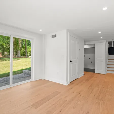 Empty bright room with light wood floors, white walls, sliding glass door, and stairs leading to kitchen area.